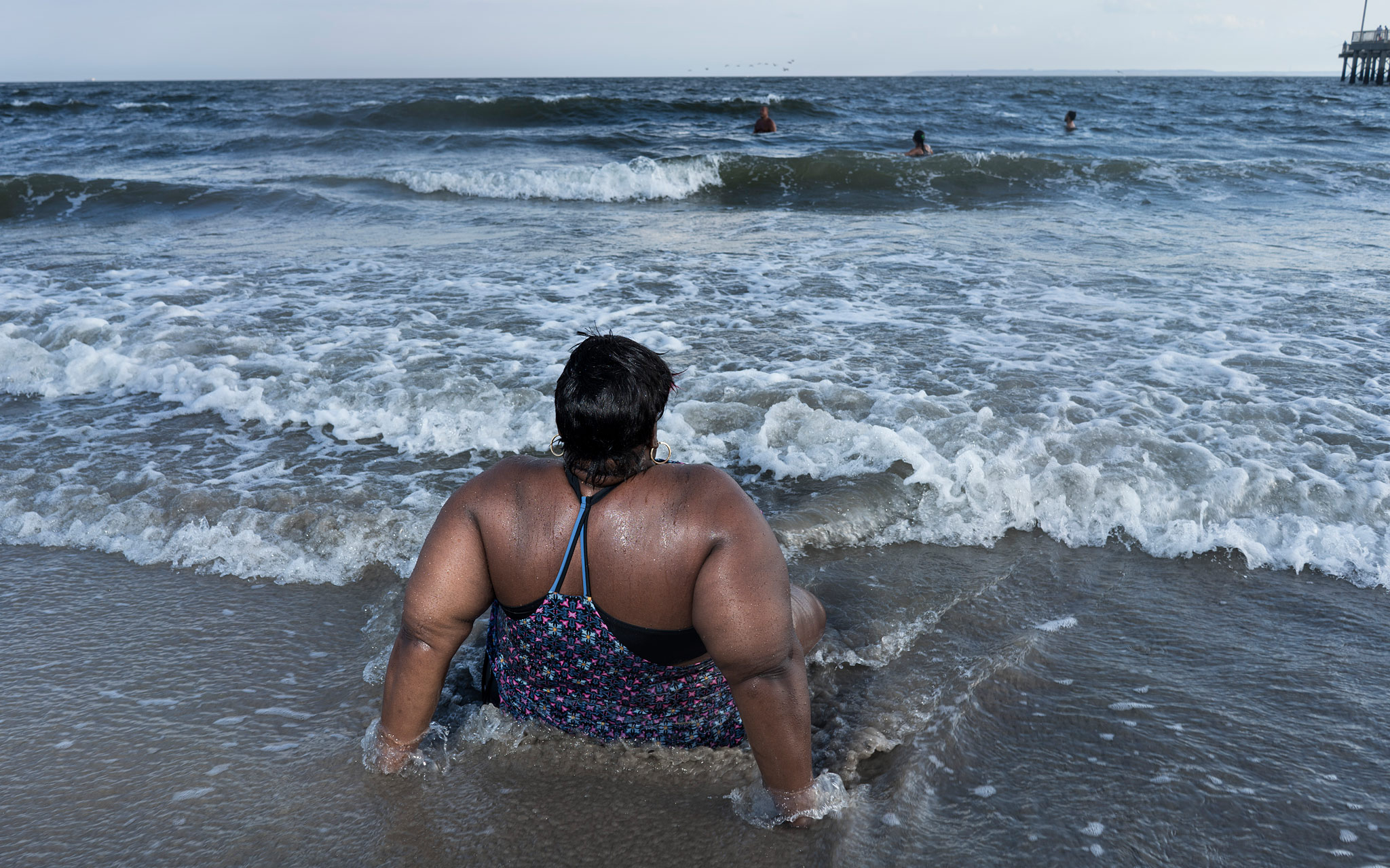 Nainen istuu rannalla puolittain vedessä. Aallot tulevat hänen jalkojensa päälle.