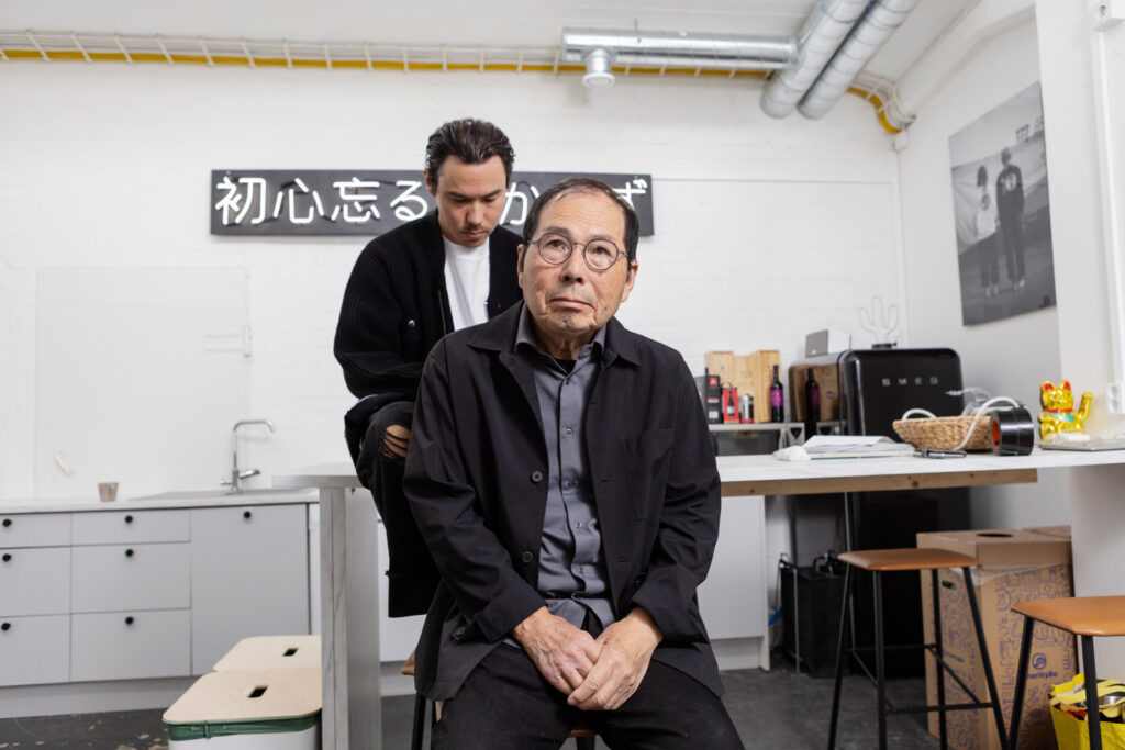 Vanhempi mies istuu nuoremman edessä työstudiossa. Miesten takana on japaninkielinen kyltti.