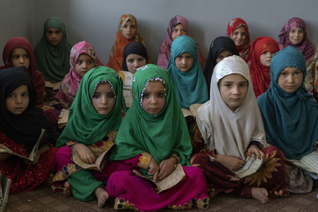 Värikkäisiin vaatteisiin ja hijabiin puetut nuoret tytöt istuvat rivissä ja katsovat vakavasti kameraan.
