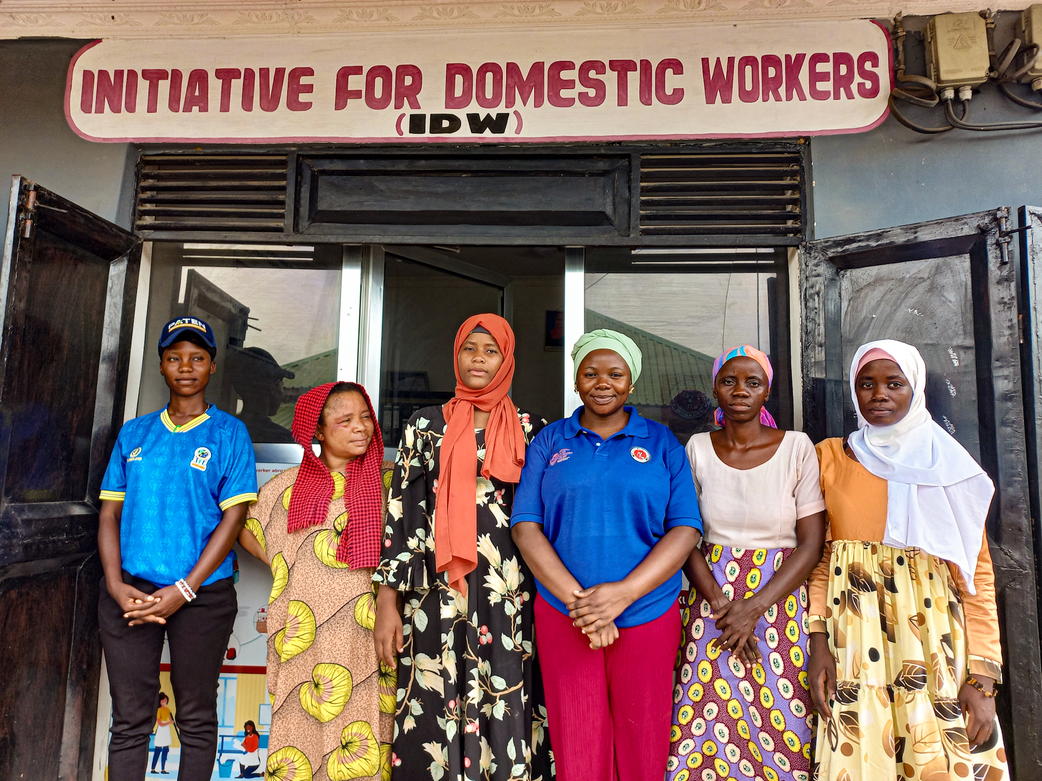 Ryhmäkuva viidestä naisesta ja yhdestä miehestä toimiston edessä, kyltissä teksti Initiative for Domestic Workers.